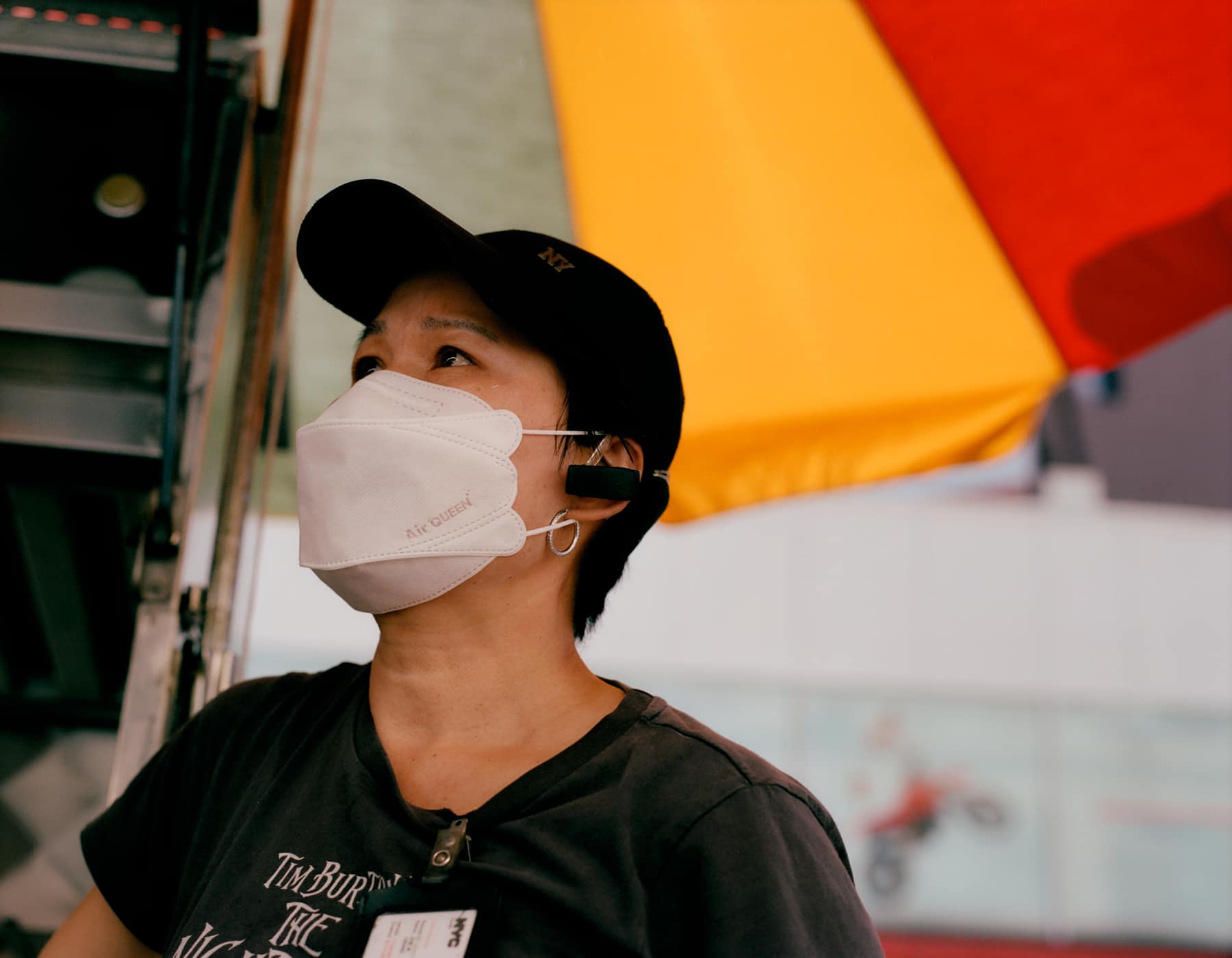 A vendor in a cap and mask under a red and yellow umbrella