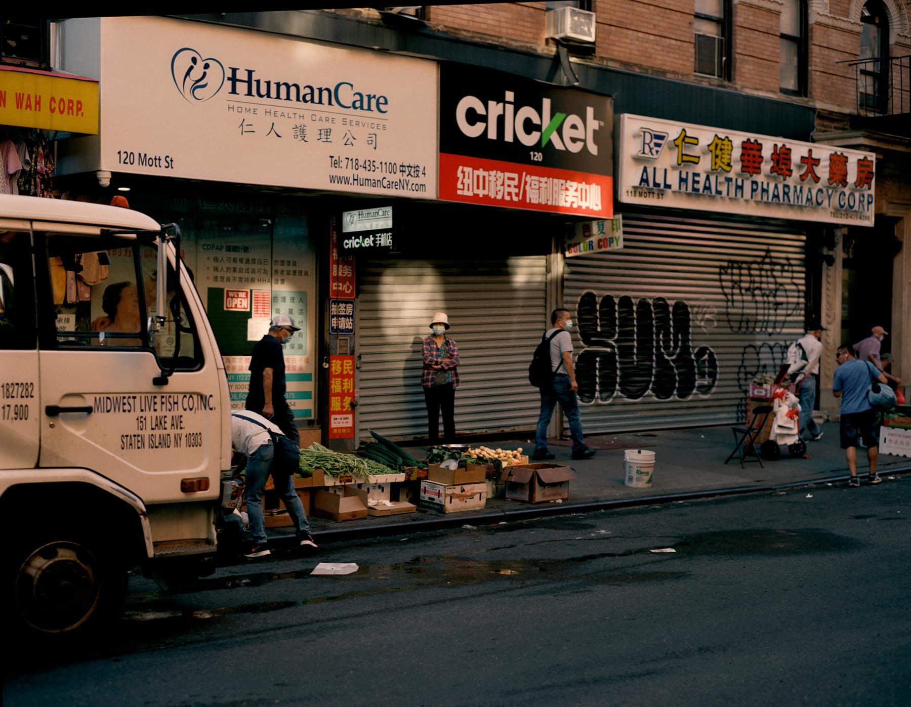 A row of Mott Street storefronts, a delivery truck and a produce stand on the sidewalk
