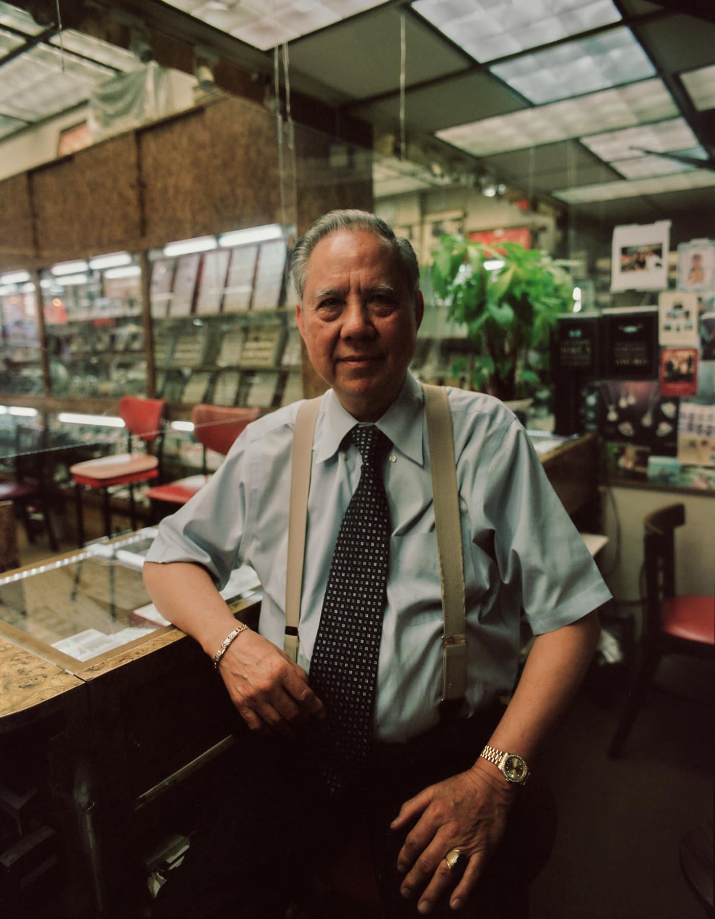 A shopkeeper in a tie and suspenders at the desk of his store, plants and a mirrored ceiling behind him