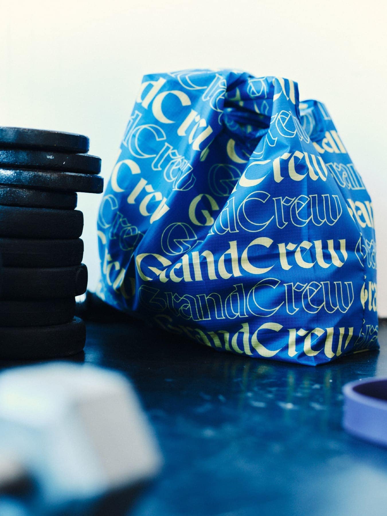 The blue Grand Crew tote bag beside a stack of weight plates