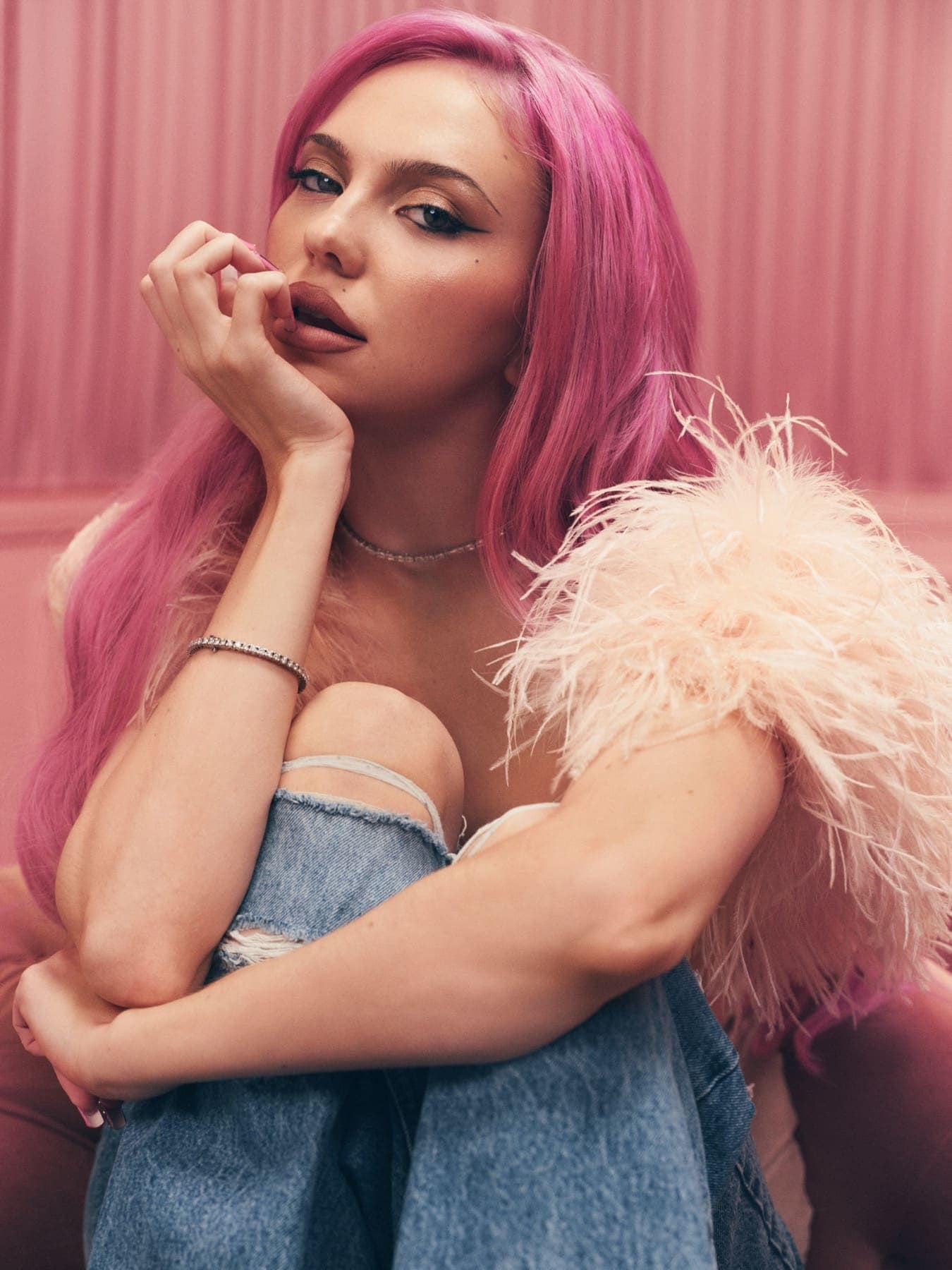 Portrait of ADÉLA in a feathered top, hand to her lips, pink hair against pink curtains