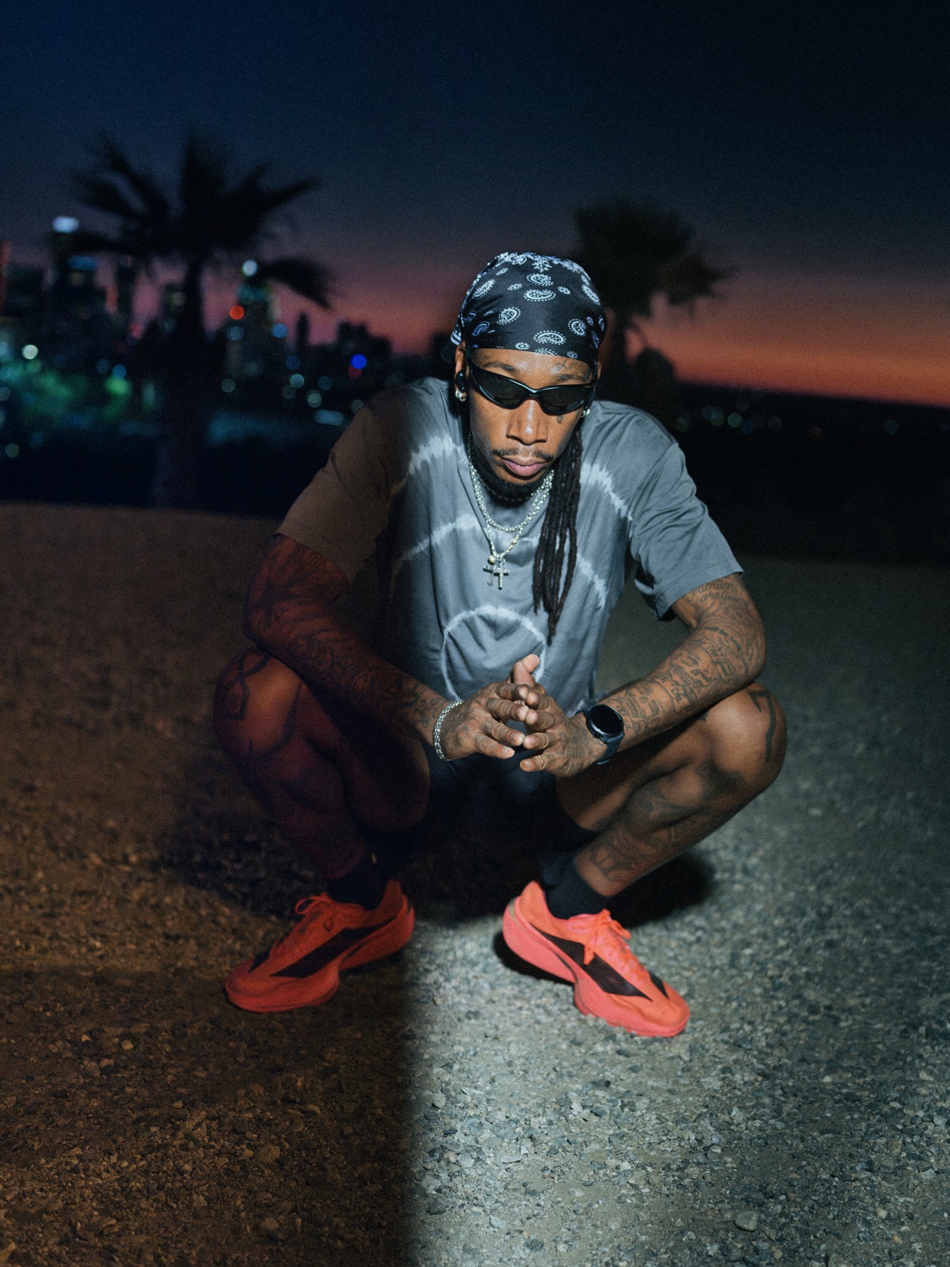 Wiz Khalifa crouched on a road at dusk, palm trees and the skyline behind