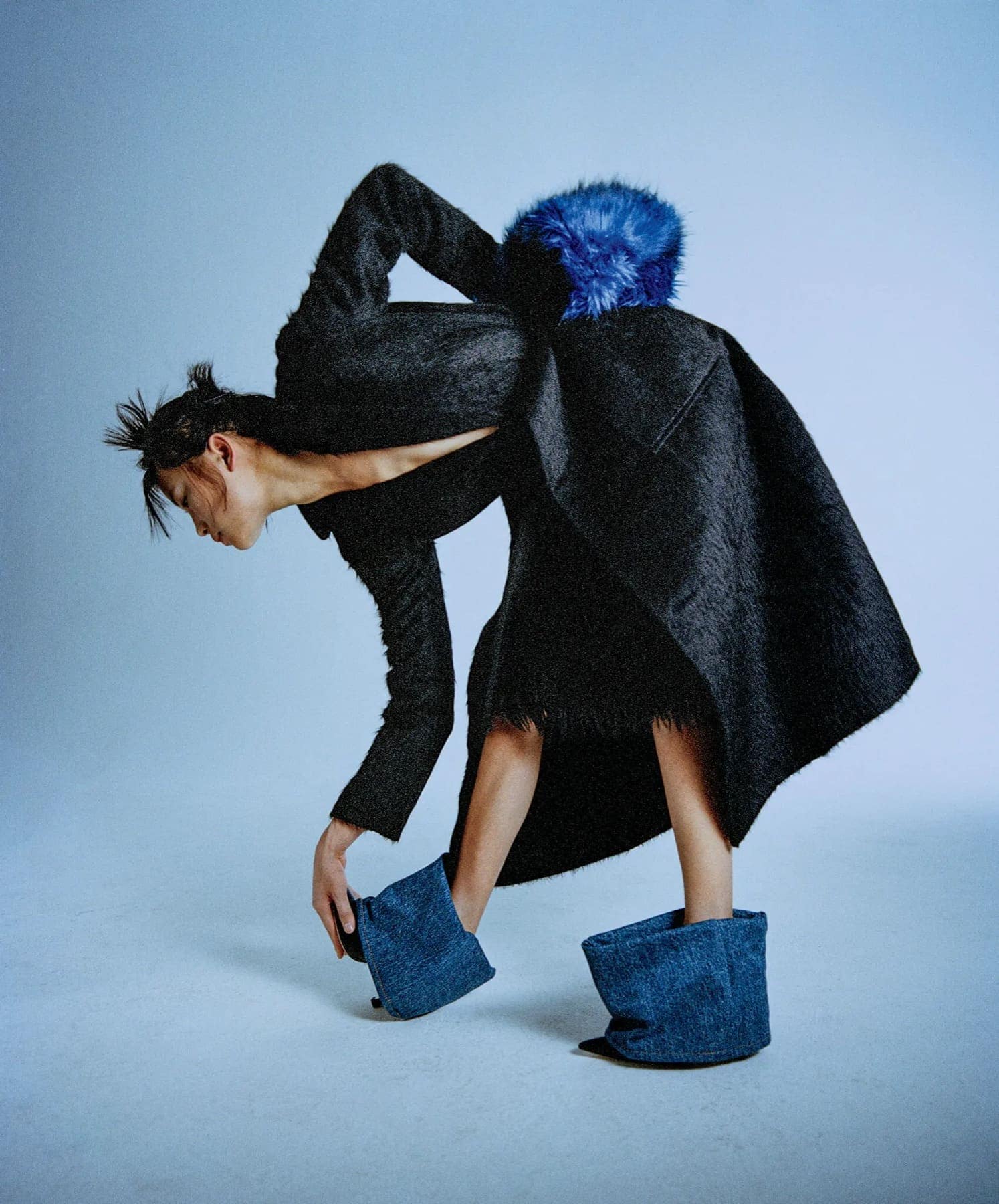 Model bent forward in a dark textured coat with a blue fur shoulder piece and blue suede platform mules
