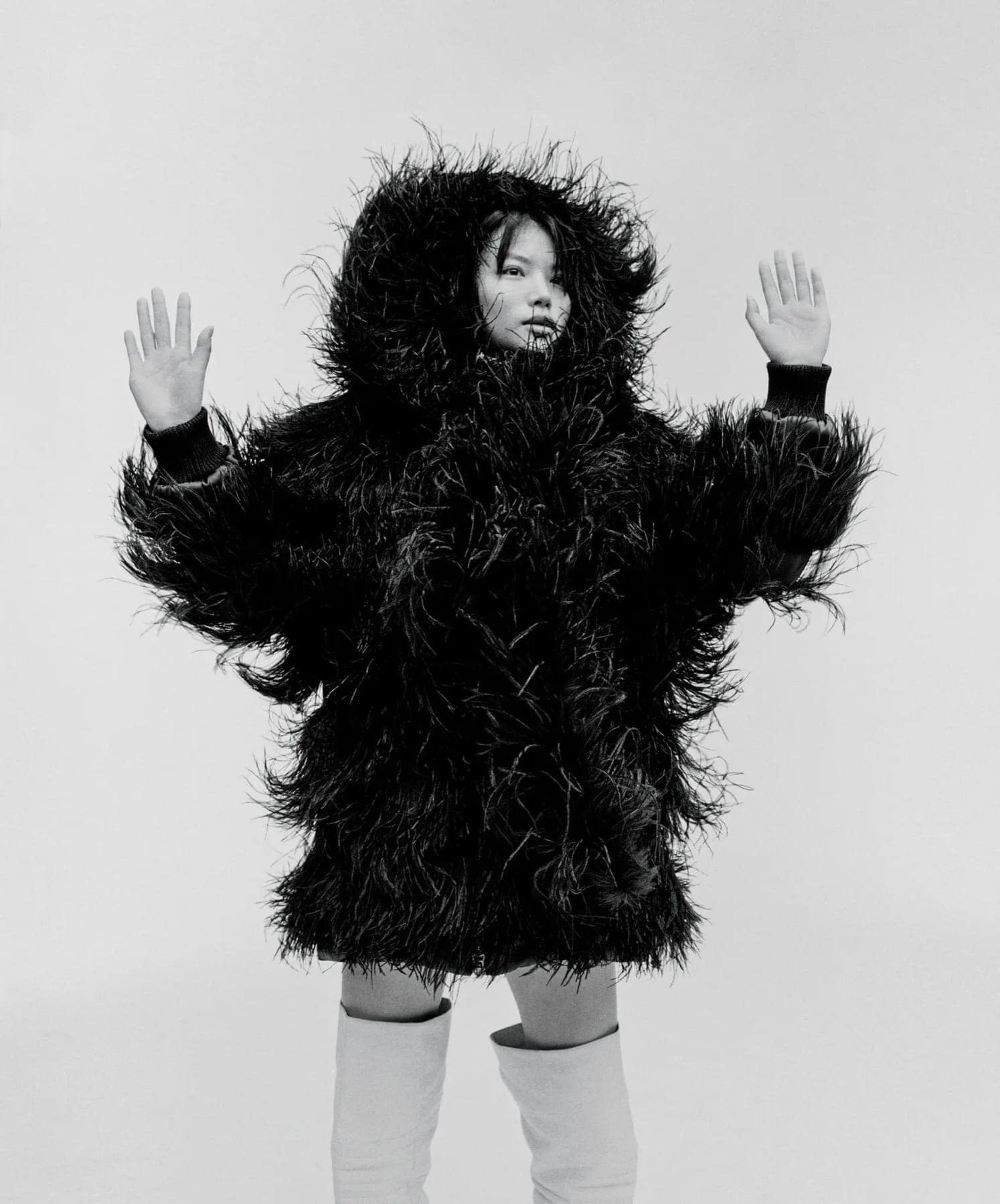 Model with arms raised in a shaggy black fur coat and hood, white boots against a pale backdrop