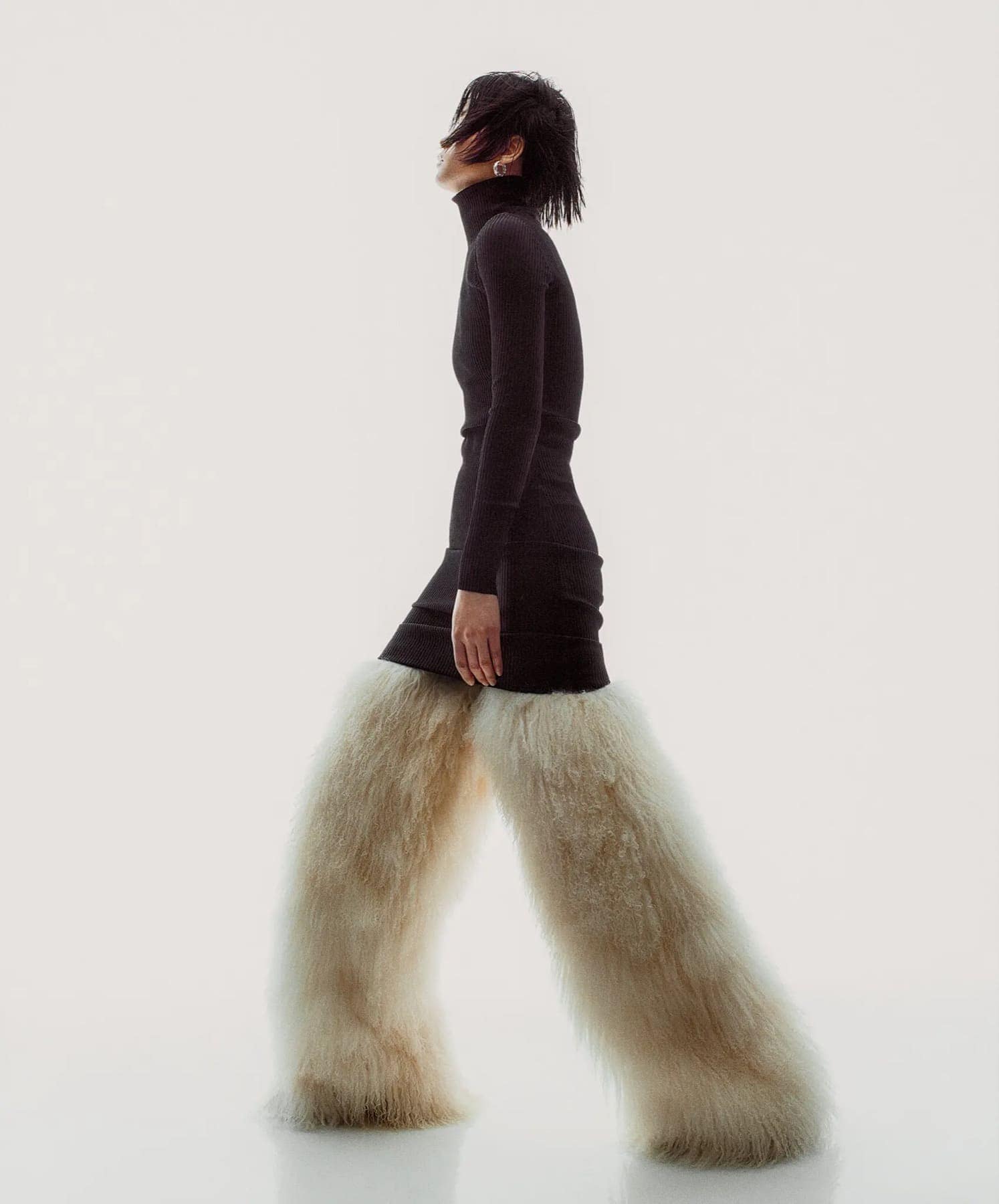 Model walking in profile in a dark ribbed turtleneck over wide cream fur-trimmed trousers