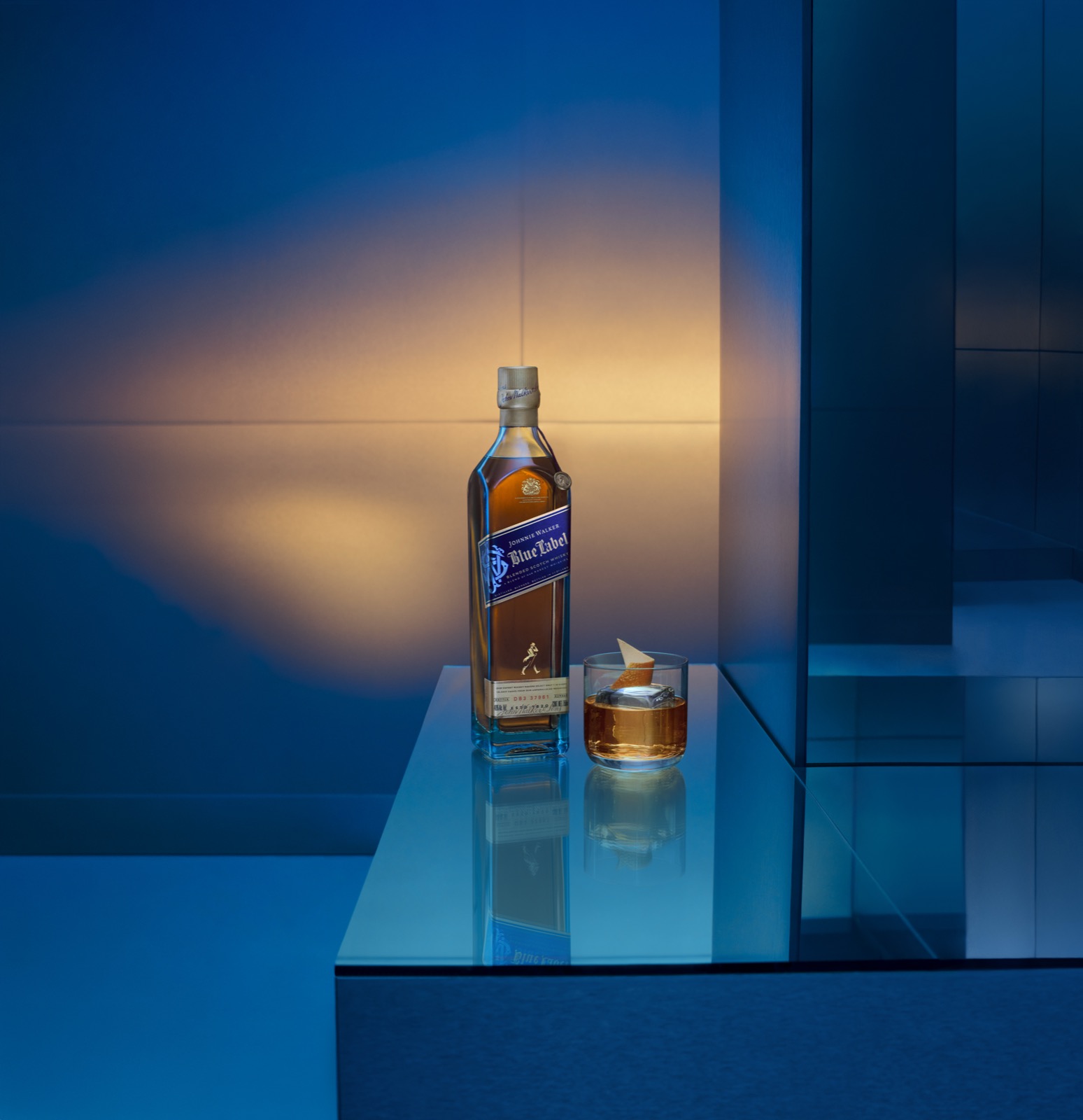 A bottle of Johnnie Walker Blue Label and a coupe glass on a mirrored table against blue architectural glass