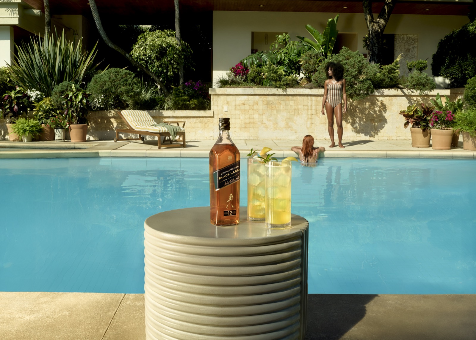 A woman descending garden steps toward a poolside table set with a highball